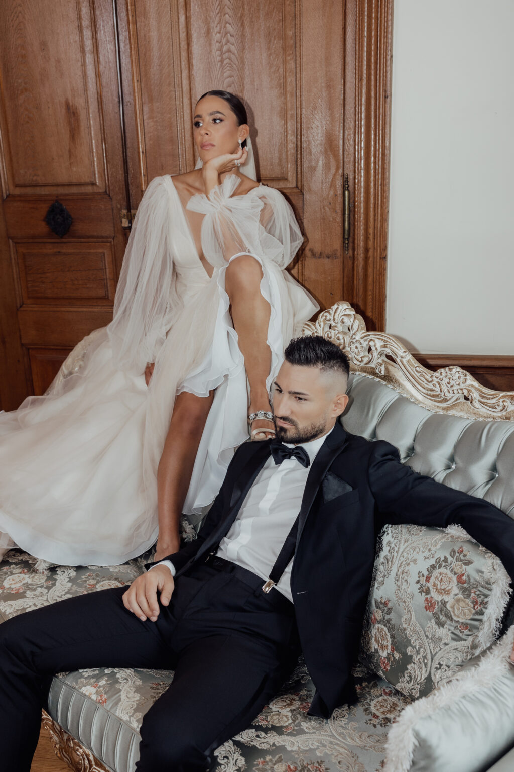 Eine Frau in einem weißen Kleid posiert mit einem Bein auf einem Sofa, während ein Mann in einem schwarzen Smoking entspannt neben ihr sitzt.