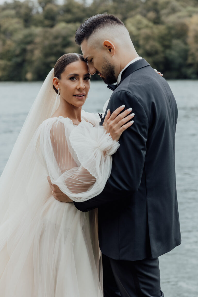 Braut und Bräutigam umarmen sich an einem See, der Bräutigam küsst die Stirn der Braut, während sie in die Kamera schaut.