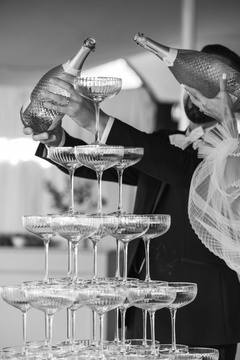 Zwei Personen gießen bei einer Feier Champagner in einen kaskadenartigen Turm aus Coupe-Gläsern.