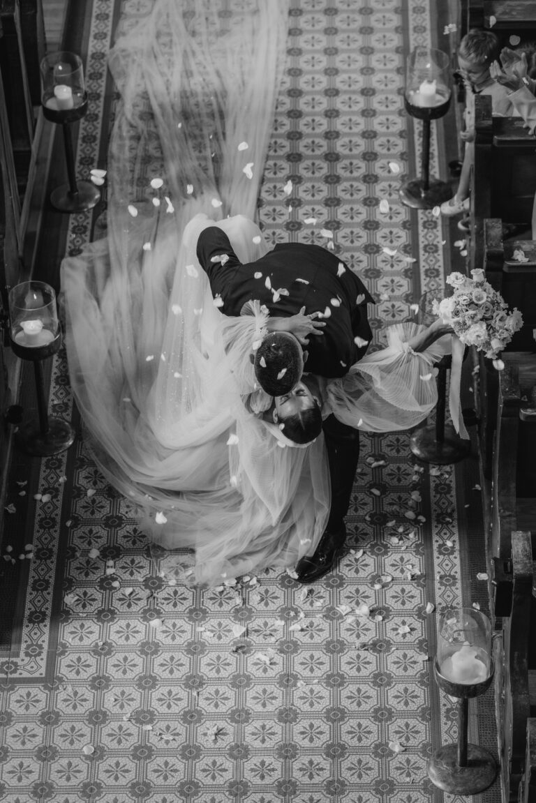 Eine Braut und ein Bräutigam küssen sich leidenschaftlich in einem Kirchenschiff, während Blumenblätter um sie herum fallen.