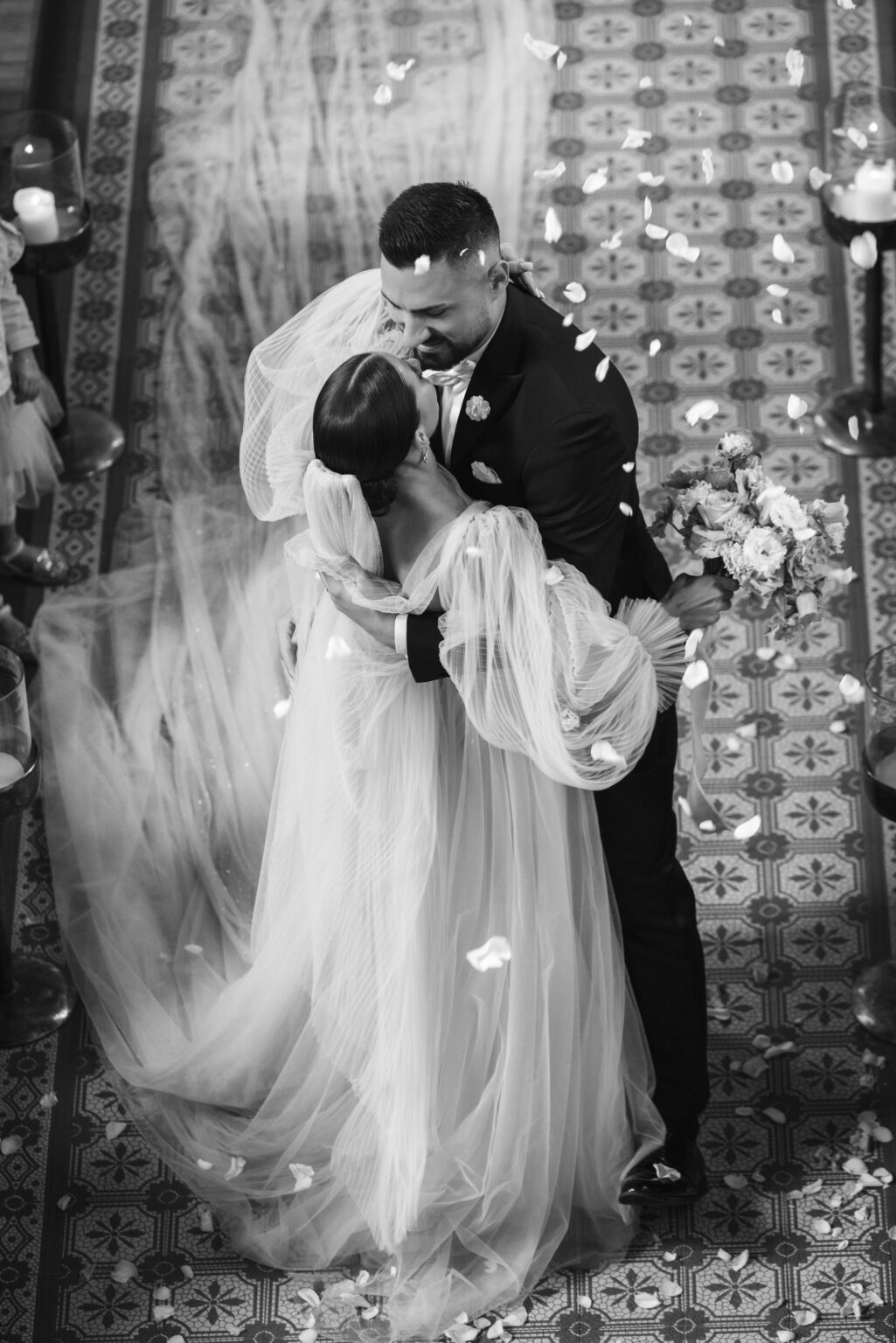 Braut und Bräutigam küssen sich am Gang, umgeben von Blumenblättern in einem wunderschön dekorierten Raum.