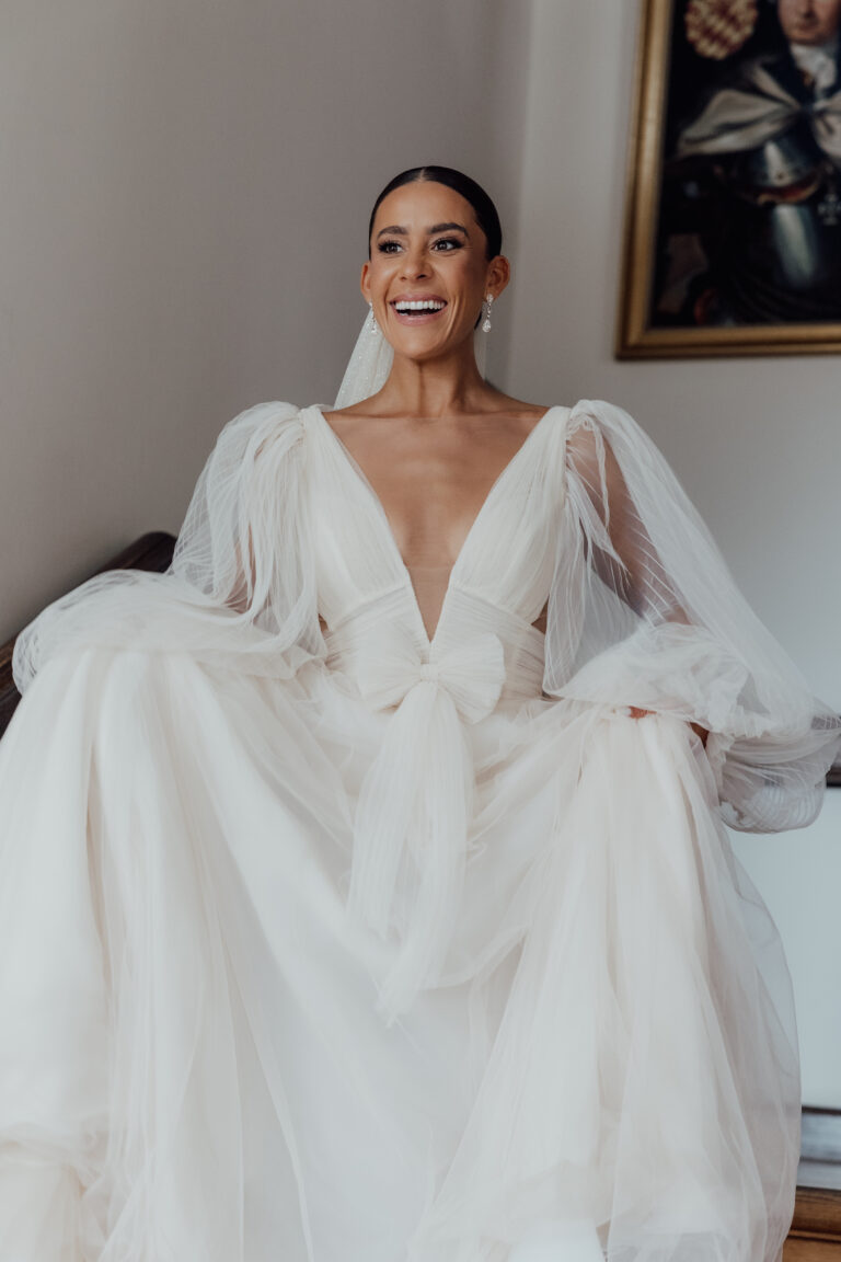 Eine lächelnde Braut in einem fließenden weißen Hochzeitskleid mit durchsichtigen, gewellten Ärmeln und einem tiefen V-Ausschnitt.