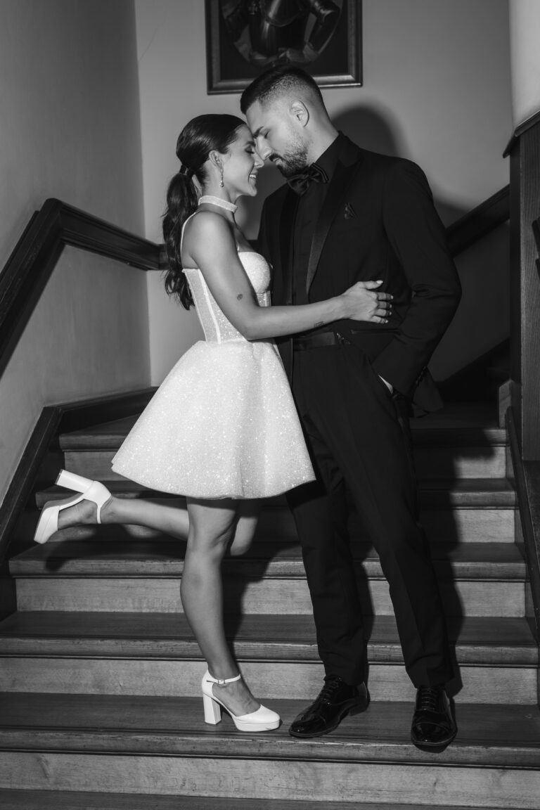 Ein förmlich gekleidetes Paar umarmt sich auf einer Treppe und lächelt sich liebevoll an.