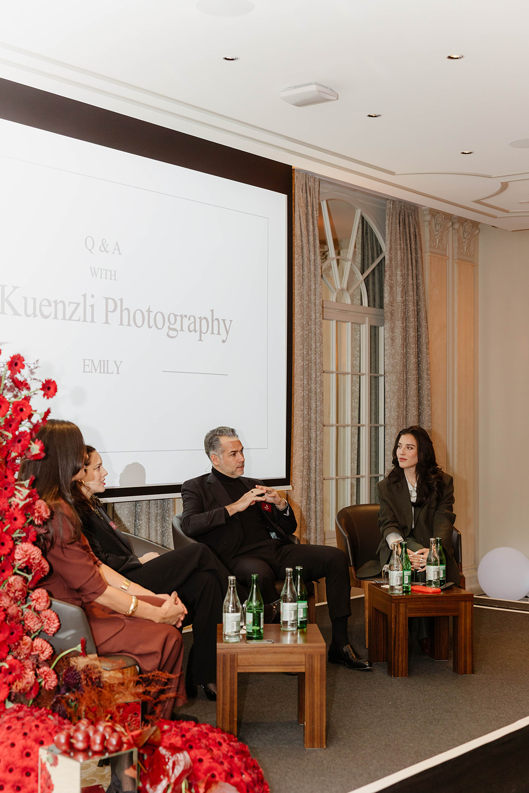 Vier Personen sitzen auf der Bühne vor einem Bildschirm für eine Fragerunde, auf den Tischen stehen rote Blumen und Wasserflaschen.