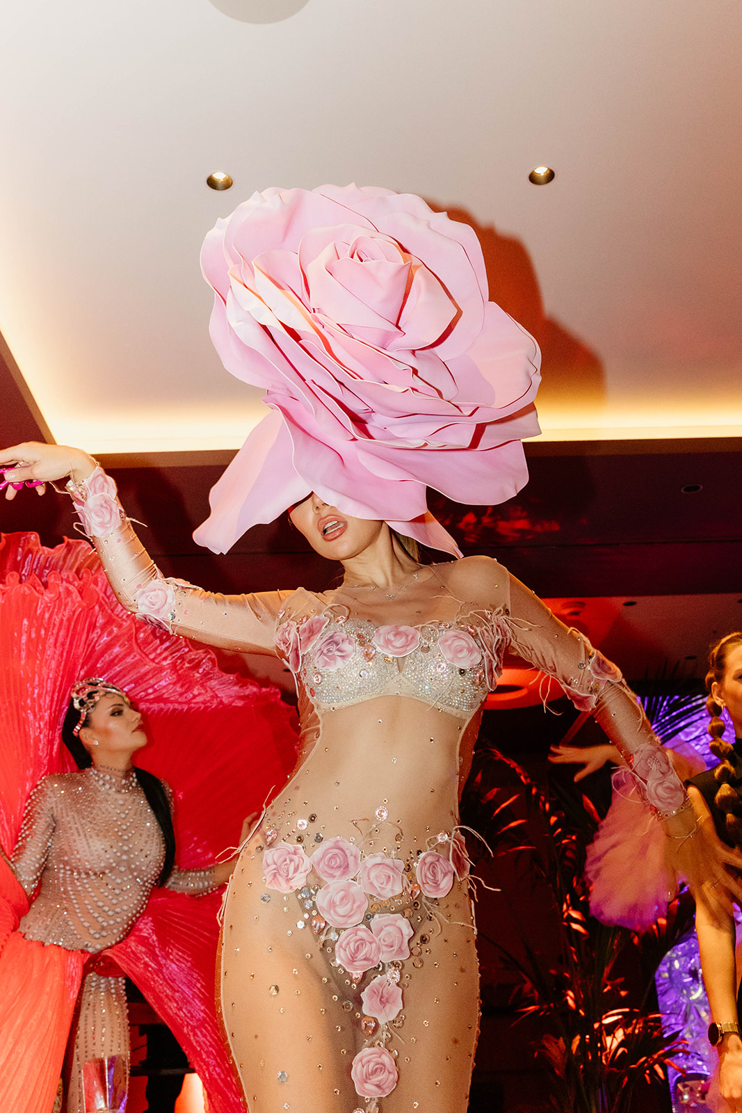 Eine Darstellerin in einem durchsichtigen Outfit mit Rosen und einem großen rosafarbenen Blumenkopfschmuck tanzt unter hellen Lichtern auf der Bühne.