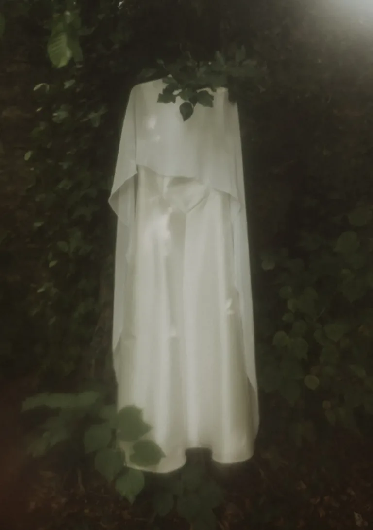 Eine weiße, geisterhafte Gestalt, in Tücher gehüllt, steht inmitten dichten grünen Laubs in sanftem, dunstigem Licht. Von Ruette-Gut am Bielersee.