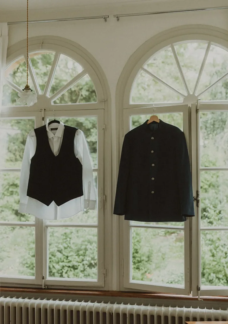 Vor großen Fenstern hängen ein weißes Hemd, eine schwarze Weste und eine schwarze Jacke mit Knöpfen. Von Ruette-Gut am Bielersee.