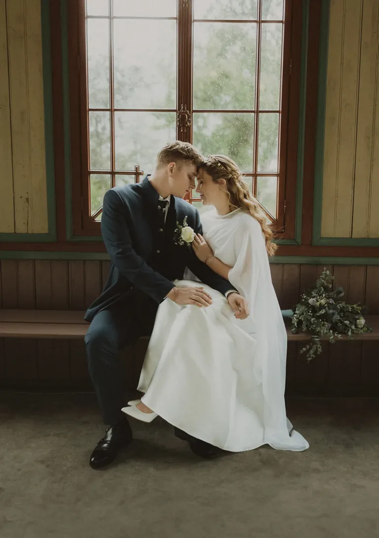 Braut und Bräutigam sitzen eng beieinander auf einer Bank am Fenster und genießen an ihrem Hochzeitstag einen intimen Moment. Von Ruette-Gut am Bielersee.