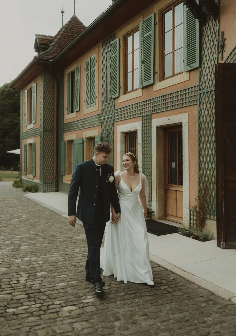 Ein Brautpaar spaziert Hand in Hand auf einer Kopfsteinpflasterstraße an einem charmanten, farbenfrohen Gebäude vorbei. Von Ruette-Gut am Bielersee.