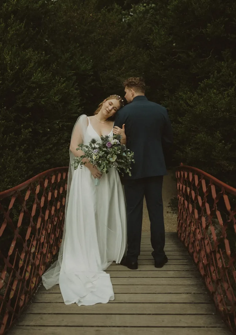 Eine Braut in einem weißen Kleid lehnt sich an ihren Bräutigam in einem dunklen Anzug, der einen Blumenstrauß hält, auf einer roten Brücke, umgeben von viel Grün. Von Ruette-Gut am Bielersee.