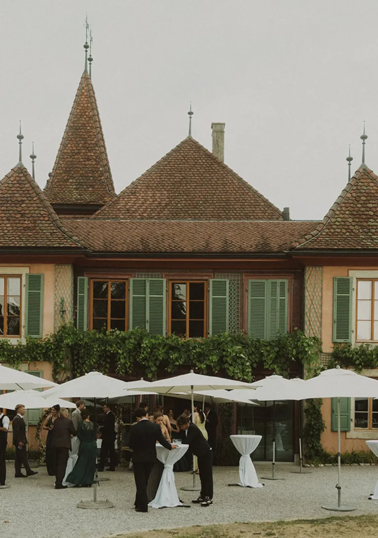 Vor einem großen, alten Gebäude mit grünen Fensterläden versammeln sich Menschen unter Sonnenschirmen und unterhalten sich. Von Ruette-Gut am Bielersee.