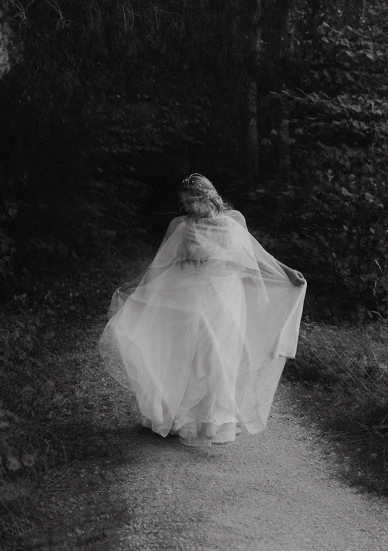 Eine Person in einem wallenden weißen Kleid geht auf einem Waldweg, umgeben von schwarz-weißen Bäumen. Von Ruette-Gut am Bielersee.