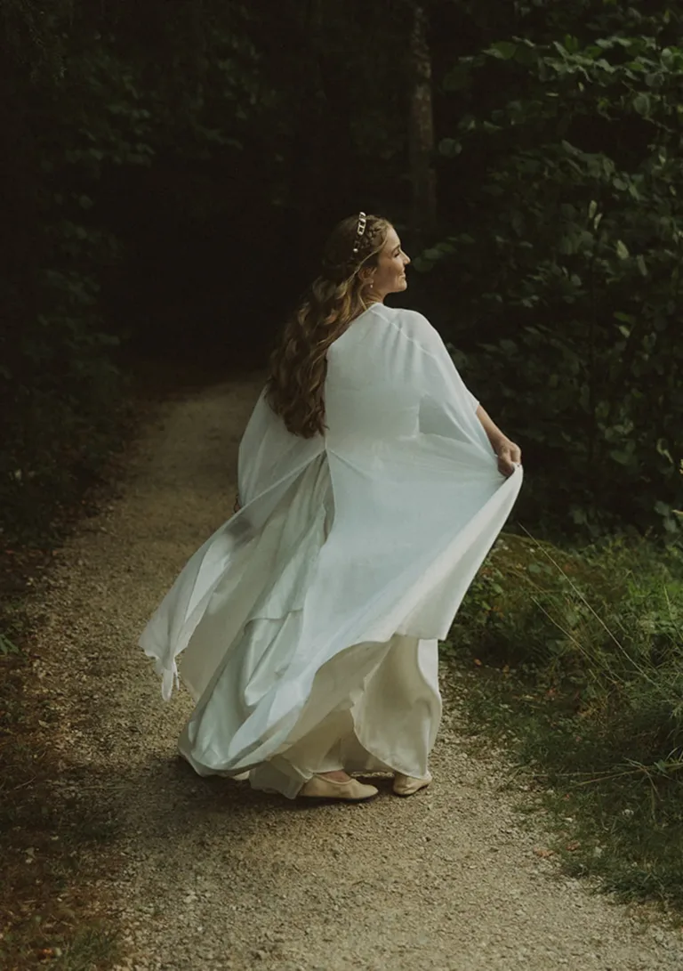 Eine Frau in einem wallenden weißen Kleid geht auf einem Waldweg, blickt zurück und lächelt, umgeben von Grün. Von Ruette-Gut am Bielersee.