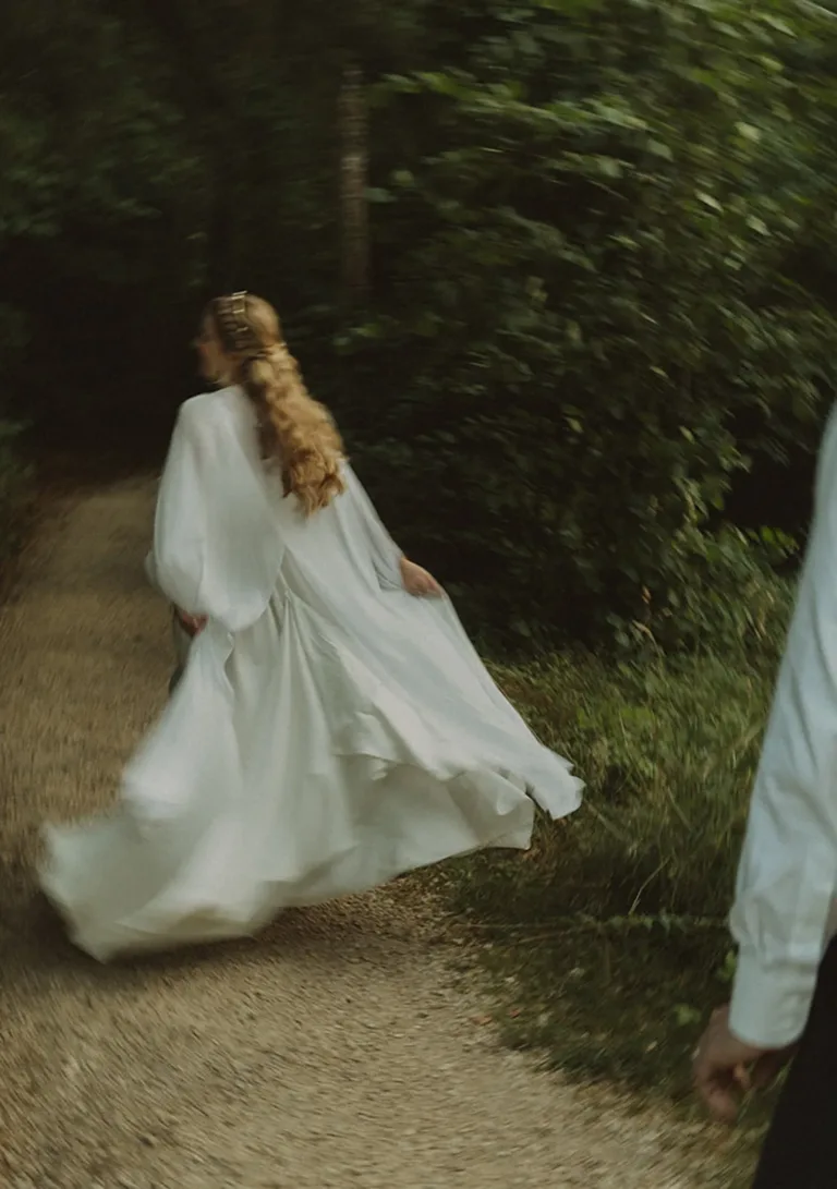Eine Frau in einem wallenden weißen Kleid läuft einen von Grün umgebenen Weg entlang, wobei eine weitere Person teilweise zu sehen ist. Von Ruette-Gut am Bielersee.