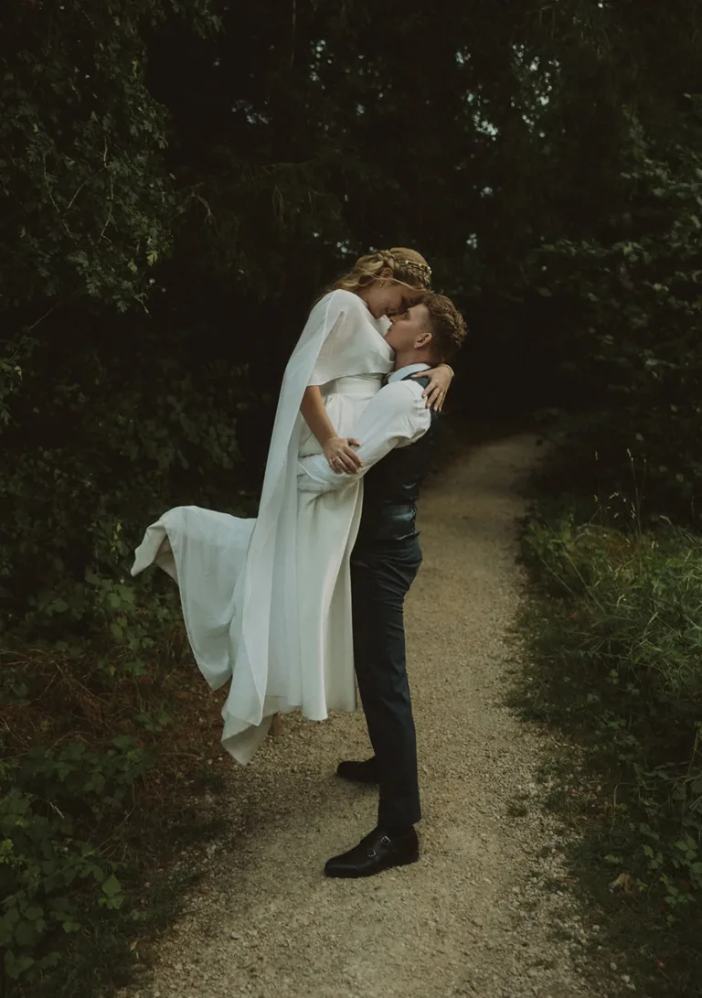 Eine Person in einem weißen Kleid wird auf einem Waldweg, umgeben von Grün, von einer anderen Person hochgehoben und geküsst. Von Ruette-Gut am Bielersee.