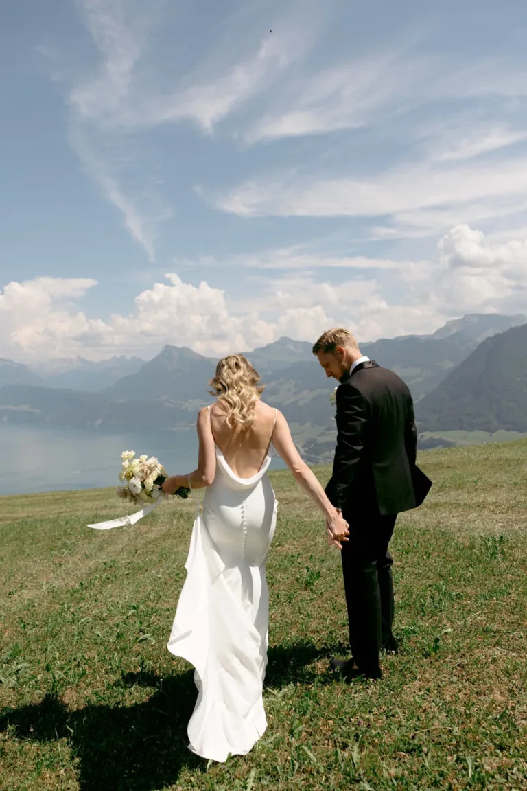 Braut und Bräutigam halten Händchen und spazieren über eine Wiese, im Hintergrund sind Berge, ein See und ein bewölkter Himmel zu sehen.
