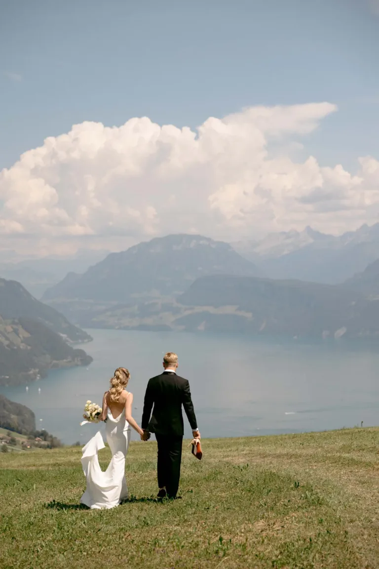 Braut und Bräutigam halten Händchen und gehen über eine Wiese auf einen See zu, im Hintergrund sind Berge und Wolken zu sehen.