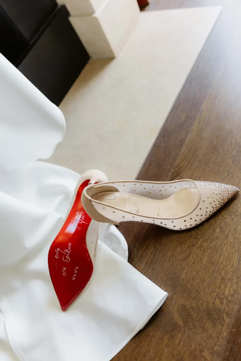 Ein mit Edelsteinen besetzter, hautfarbener High Heel mit einer Aufschrift auf der roten Sohle liegt neben einem weißen Kleid auf einem Holzboden.