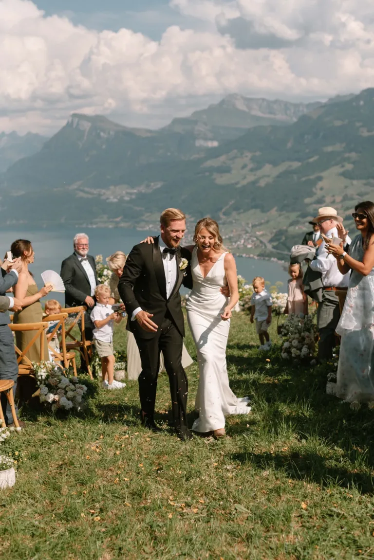 Ein frisch verheiratetes Paar schreitet freudig einen Gang im Freien entlang, umgeben von Gästen und einer Bergkulisse.