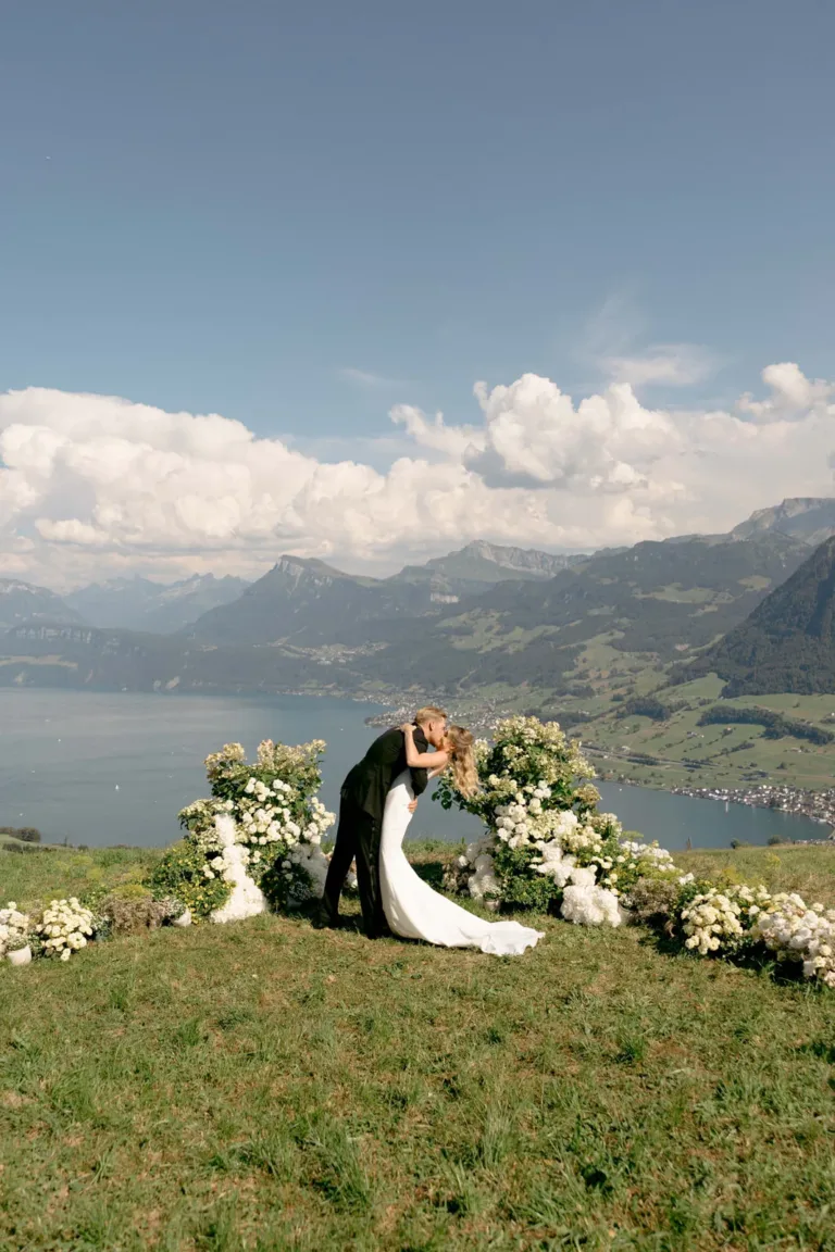 Ein Paar küsst sich bei einer Hochzeit im Freien, im Hintergrund sind Berge, ein See und Blumenarrangements zu sehen.