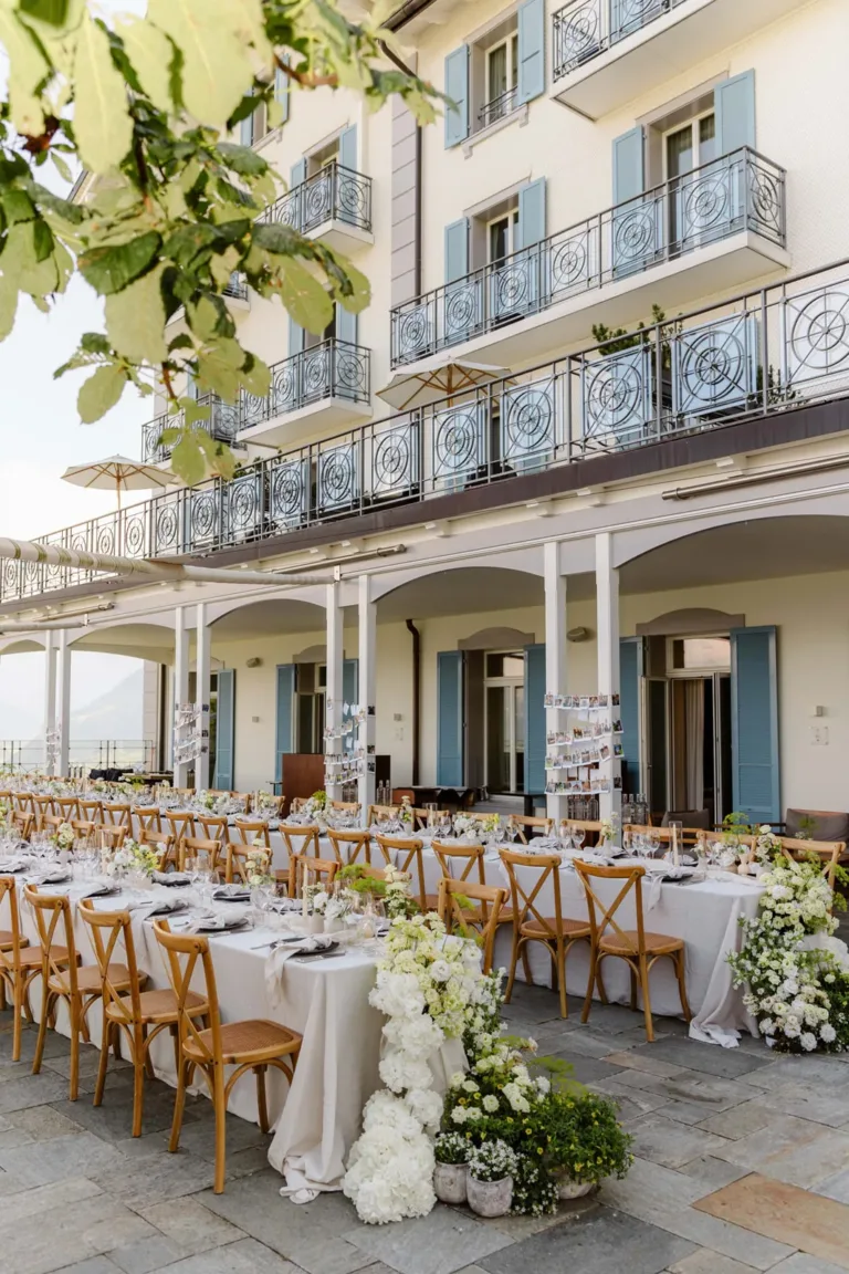Elegante Hochzeitsfeier im Freien mit langen Tischen, weißen Blumen und einem Gebäude mit blauen Fensterläden im Hintergrund.