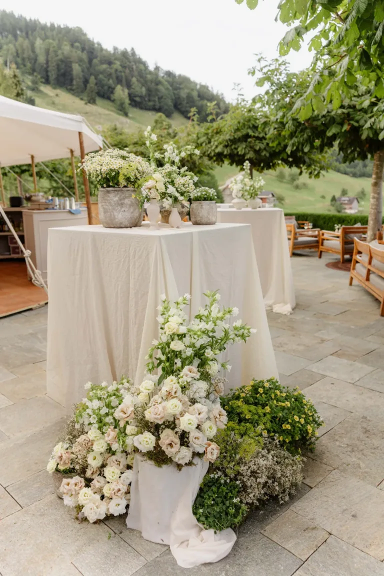 Ein hoher Tisch, mit einem weißen Tischtuch bedeckt und mit weißen und grünen Blumenarrangements geschmückt, steht im Freien auf einer Steinterrasse.