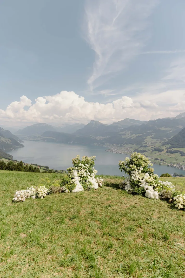 Weiße Blumenarrangements auf grünem Gras bieten unter einem teilweise bewölkten Himmel einen Blick auf einen See und die Berge.