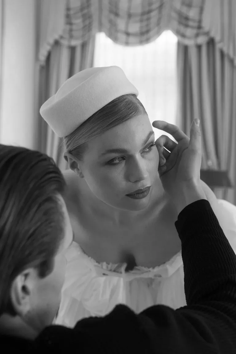 Eine Hochzeitsplanerin für Luxushochzeiten richtet den Ohrring einer Frau zurecht, die einen Pillbox-Hut trägt.