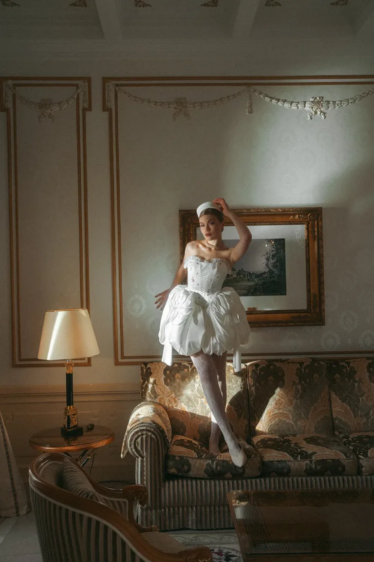 Eine Person in einem weißen Korsettkleid und einem Pillbox-Hut, die für eine Hochzeit im Ausland auf einem Sofa steht.