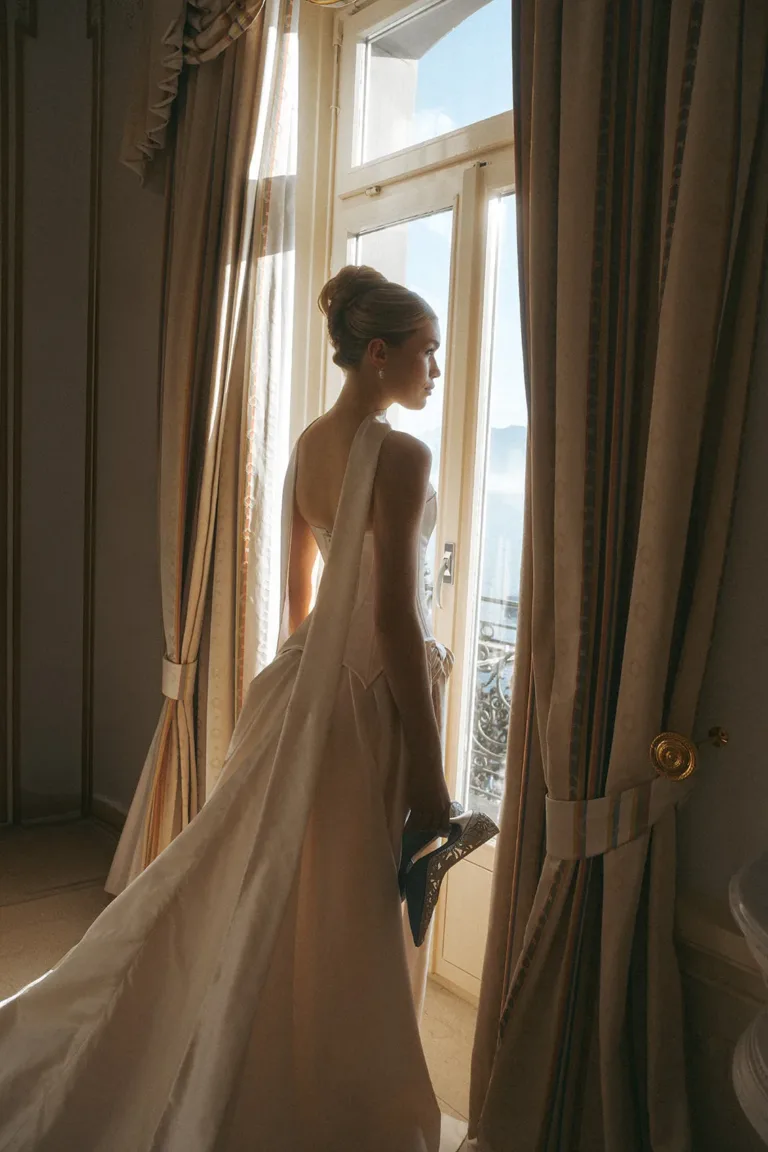 Eine Braut in einem Brautkleid steht am Fenster und hält Schuhe für ihre Hochzeit in der Schweiz in der Hand., Luxury Wedding Planner, Zurich, The WES, Switzerland, Schweiz, Lara Flueckiger, Grand Hotel National, Luzern