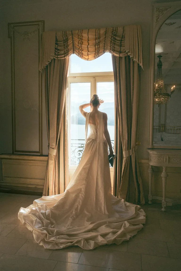 Eine Braut in einem Brautkleid steht bei ihrer Hochzeit in der Schweiz an einem Fenster mit Blick auf das Wasser, Luxury Wedding Planner, Zurich, The WES, Switzerland, Schweiz, Lara Flueckiger, Grand Hotel National, Luzern