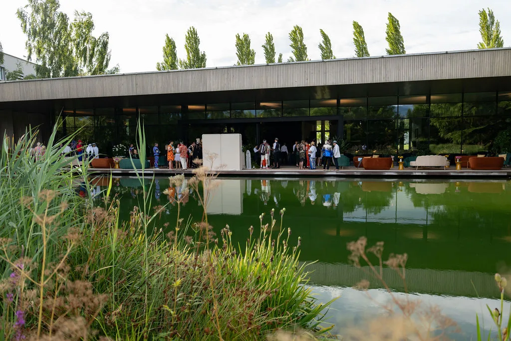 Vor einem modernen Gebäude – ideal für Firmenveranstaltungen in der Schweiz – versammeln sich Menschen, die sich in einem grünen Teich spiegeln.