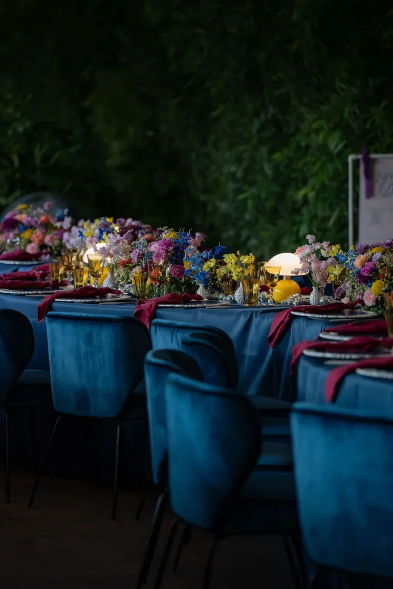 Ein eleganter Tisch für Firmenveranstaltungen in der Schweiz mit blauen Samtstühlen, farbenfrohen Blumen und leuchtenden Lampen in der Abenddämmerung.