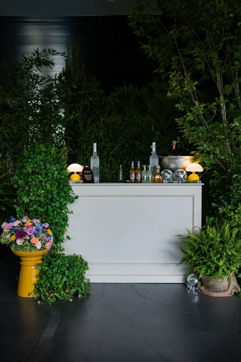 Eine weiße Theke von Corporate Events Schweiz, umgeben von grünen Pflanzen, Flaschen, Blumen auf einem gelben Hocker und kleinen Lichtern.